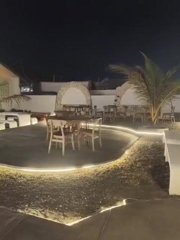The video shows an outdoor seating area of a restaurant or cafe at night. The area is lit by strings of lights and spotlights. There are several tables and chairs set up, some with umbrellas. A palm tree is visible in the center of the scene. The ground is a mix of dark gravel and concrete. In the background, there is a building with arched windows. There are a few people walking around in the background.