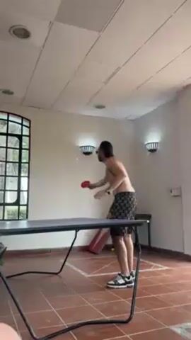 A shirtless man in dark printed shorts and black-and-white sneakers plays ping pong in a tiled room. The room has a large window, modern wall sconces, and a tiled ceiling with recessed lighting. The man serves the ping pong ball, misses the table, and grimaces. A child's slide is visible in the background.