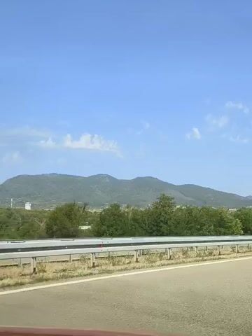 The video shows a view from the inside of a moving vehicle on a highway. The road is asphalt with white lane markings. To the left of the road, there is a metal guardrail, followed by a grassy area with trees and then distant hills and mountains under a blue sky with scattered clouds. A few buildings are visible in the distance on the left side. A dark-colored car is traveling ahead in the same direction on the highway. The video appears to be filmed during the daytime, suggesting sunny weather.