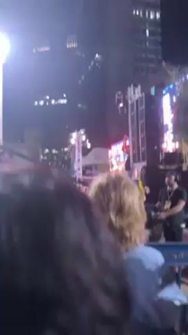 The video shows a gathering at night in an outdoor space with a stage. The main subject in the frame is the back of a person with blonde hair. Behind them, a stage is visible with an illuminated screen showing a countdown timer. Two people are seen standing near the stage holding signs. A man in a black shirt and cap stands by the side of the stage, possibly operating equipment. The background includes buildings and other crowd members. The atmosphere appears to be one of protest or demonstration, indicated by the signs and the gathering itself.
