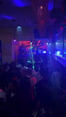 The video shows a crowded room with people dancing.  The room is dimly lit with purple, blue and green lights, and there appear to be lights projecting beams of color toward the crowd. A DJ is positioned in a booth above the crowd, with the lights shining on them. There are what appear to be tropical decorations hanging from the ceiling and on the walls. The video appears to be shot from within the crowd, looking up at the DJ booth.