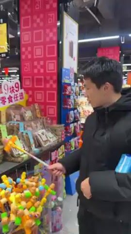 A man in a black jacket is standing in a grocery store aisle. He is holding a yellow and silver toy hammer and hitting it against a metal shelf. The shelf is filled with packages of snacks. There is a sign above the snacks that says "9块9 零食任选" in Chinese characters. In front of the shelf is a basket filled with toy hammers, and the man is standing in front of that basket. The store is well-lit and decorated with red and white patterns.