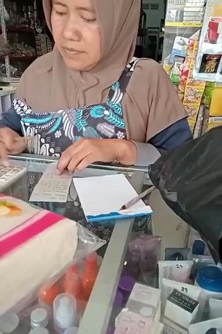 A woman wearing a light brown hijab and a brown shirt with a patterned bib is standing behind a counter. She is looking down at a calculator and a receipt. Her right hand is on the calculator, and her left hand is pointing at the receipt. Next to the receipt is a small notepad with a pen resting on it. In front of her, on the counter, are some packaged goods, including what appears to be food items and various bottles and boxes of cosmetics. A black plastic bag is partially visible on the right side of the counter. The background shows shelves stocked with various products. The lighting appears to be indoor. The woman seems to be in the process of totaling an order or checking prices.