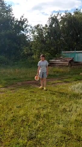 A young girl, appearing to be in her pre-teen years, is playing soccer in a grassy outdoor area. She is wearing a light blue t-shirt and denim shorts, with yellow shoes. She is actively kicking a multi-colored soccer ball with her right foot. The setting appears to be a rural area, possibly a backyard or a field, with lush green grass and trees in the background. There are also some wooden planks stacked up and a light blue fence visible behind the girl. The sky is partly cloudy, suggesting it might be daytime, perhaps late afternoon given the lighting. The girl seems engaged and focused on her activity, with a neutral or slightly concentrated expression. The overall atmosphere is casual and leisurely, indicating a moment of outdoor recreation. The context provided places this scene in Tashkazgan, Russia. There is no visible text in the media file.