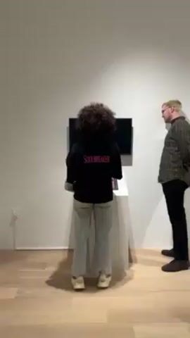Two people are standing in front of a piece of art. The person on the left is wearing a black shirt with the word "SOULBREAKER" in pink letters on the back, light-colored pants, and gold sneakers. They have curly hair. They are interacting with a touch screen on a white pedestal. A black flat screen monitor is mounted on the wall behind the pedestal. The person on the right is wearing a plaid shirt, black pants, and black shoes. They have short, bleached blonde hair and glasses. They are looking at the person on the left and the touch screen. The room appears to be an art gallery with white walls and light wood floors. Other people walk by in the foreground.