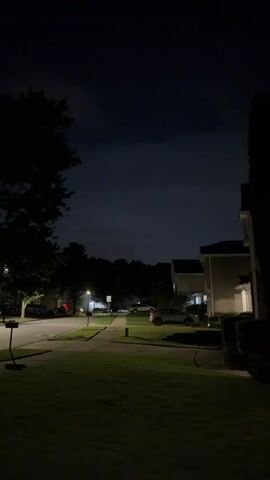 The media file captures a quiet residential street at night, likely in Buford, United States. The scene is dimly lit by streetlights and the ambient glow from house windows, casting long shadows across manicured lawns and sidewalks. Several houses, typical of a suburban neighborhood, line the street, with cars parked in driveways and on the roadside. Trees frame the edges of the image, adding to the sense of a peaceful, secluded environment.

The most striking element of the scene is a faint lightning flash illuminating the dark, cloudy sky in the distance. This suggests an ongoing thunderstorm, adding a touch of dramatic atmosphere to the otherwise tranquil setting. The time of day is clearly nighttime, emphasized by the darkness and artificial lighting. The weather is stormy, with the lightning indicating active thunder.

No people are visible in the frame, contributing to the feeling of stillness and solitude. The focus is on the architecture, the natural elements of trees and lawn, and the subtle evidence of weather. The overall mood is serene, with a hint of underlying power due to the distant storm. There is no visible text within the media file.