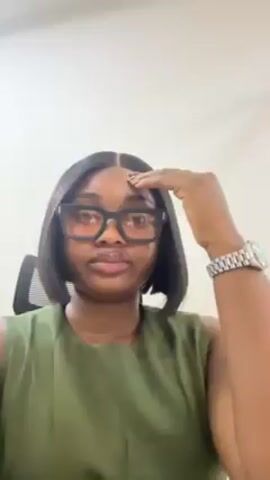 A woman with shoulder-length black hair is centered and looking directly at the camera. She wears dark-rimmed square glasses, a sleeveless olive green top, and a silver watch on her left wrist. Her left hand is raised towards her forehead, touching her hair, as if to check her hairline. She sits in an office chair against a plain white wall. The lighting appears to be bright and even.