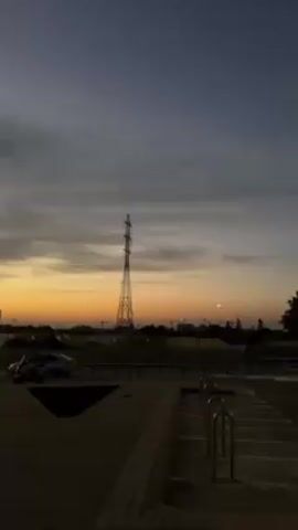 The video shows a view from an elevated position of a parking lot during a sunset. The sky transitions from dark gray at the top to a vivid orange on the horizon. There are multiple cars parked in the lot, mostly dark-colored. Tall street lights illuminate the parking area. In the distance, an electrical transmission tower is visible against the sunset. The camera moves slightly, panning across the parking lot.