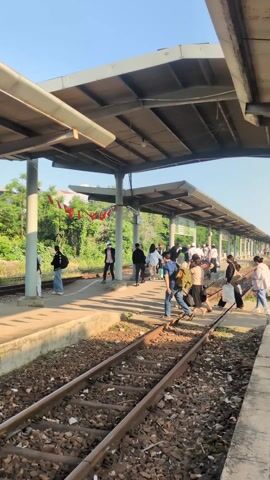 The video shows a train platform with several people walking across the tracks. They are traveling from one side of the platform to the other. The platform is covered by a metal roof with support beams. There are trees and other vegetation visible in the background. The sky is clear and blue. A speaker is making an announcement about train SE7.