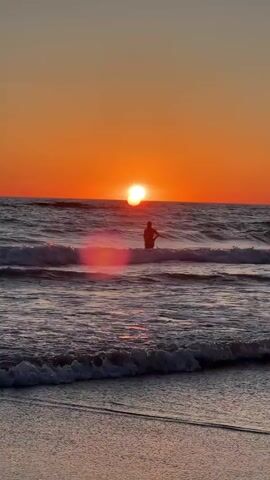 A lone silhouette of a person stands in the ocean water as waves crash around them during sunset. The sun is a bright orange orb setting on the horizon, casting a warm glow over the water and sky. The person appears to be facing the sun, possibly surfing or enjoying the view. The water is choppy with white foam from the waves. The beach is visible in the foreground, appearing wet and reflecting the sunset colors.