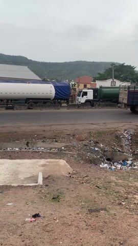 The video depicts a roadside scene in a somewhat rural or developing area. The foreground is characterized by a dirt and gravel surface with scattered trash and a concrete drainage ditch filled with debris. A motorcycle is parked in the lower center of the frame. Along the road are several large trucks, including tanker trucks, in a line, with traffic moving intermittently. In the background, there are houses, trees, and a hill or mountain range under an overcast sky. A makeshift shelter or stall is visible under some power lines with a person sitting inside. Other vehicles such as a yellow auto rickshaw and a red car are present on the road. The overall impression is one of a busy but somewhat unkempt roadside environment.
