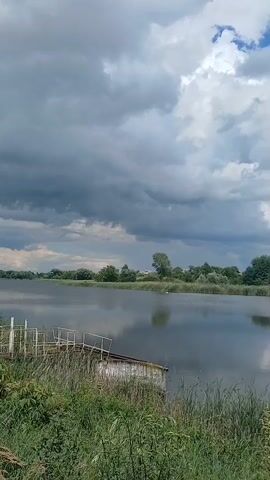 The video shows a body of water with a wooden pier and reeds in the foreground. The sky is cloudy, with some patches of blue visible. The trees on the opposite bank are green and lush. There is a small structure on the pier, possibly a boat house. The water is calm and reflects the sky and surrounding vegetation. The overall impression is one of a peaceful, natural landscape.