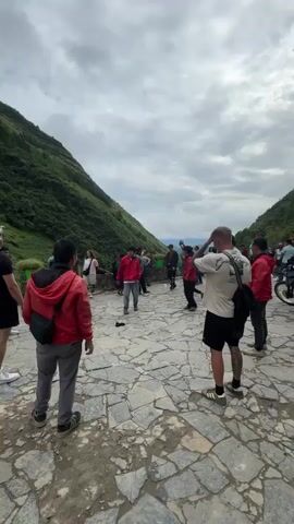 The video shows a group of people in a stone-paved area between two steep green hills under a cloudy sky. Many are wearing red jackets. Children are running around. One child in a pink dress and another in green are particularly active. A few men are standing with their backs to the camera, observing the scene. One man is kicking something, possibly a stone. The overall atmosphere seems lively and perhaps festive. A motorcycle is parked on the right side of the frame.
