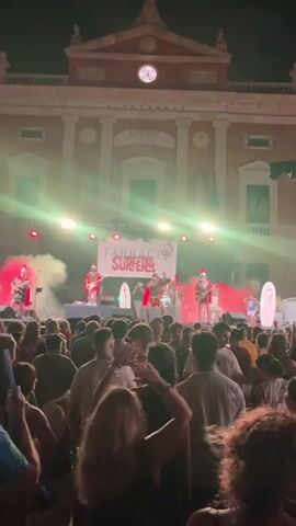 A crowded outdoor concert at night. A band performs on a stage in front of a large building. The band members are dressed in Roman-style costumes, complete with helmets and red capes. One member plays a guitar, another sings into a microphone, and another plays drums. Smoke and colored lights emanate from the stage. A large banner behind the band reads "TARRACO SURFERS". The audience is a dense crowd of people, many with their hands raised in the air. The lighting is dim, with bright spotlights on the stage.