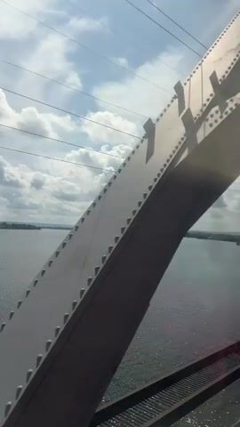 The video captures a view from a bridge overlooking a wide body of water under a cloudy sky. Metal structural elements of the bridge, including beams and railings, are visible in the foreground, framing the scene. Several power lines are strung across the sky. The water reflects the light, creating a shimmering effect. In the distance, the shore is visible, with some landmasses and possibly structures. The perspective suggests movement, as if the camera is traversing the bridge. There are no people or animals visible.