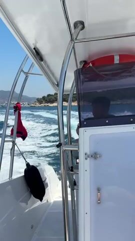 The video is filmed from the perspective of someone on a moving boat. The boat is white and has a metal frame with a white canopy. A red life vest is hanging from the frame, and a black fender is attached to the side of the boat. In the foreground, part of the boat's interior is visible, including a windshield and a person sitting at the helm. The person's face is reflected in the windshield, and they appear to be wearing a dark shirt. In the background, the ocean is choppy with white wakes trailing behind the boat. There are green islands in the distance with some buildings and trees. Another, larger boat is visible further away. The sky is clear and blue. The camera pans slightly, showing the movement of the boat and the surrounding environment.
