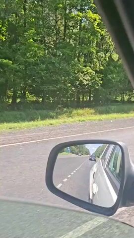 A car is driving down a paved road. Trees line the side of the road, and the sky is visible above the trees. The camera is inside the car, looking out the side window at the passing scenery. A side-view mirror reflects the road behind the car, showing other cars driving in the distance.