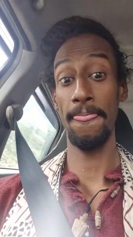 A man with dark skin and curly hair is inside a car. He is wearing a maroon jacket with a patterned scarf. The man is looking at the camera with a slight smirk. Through the car window, a mountainous landscape with patches of snow is visible. The overall scene appears to be a road trip through a scenic, possibly remote, location.