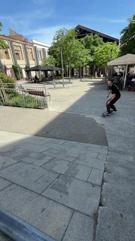 A person on a skateboard rolls down a concrete slope outdoors on a sunny day. The skateboarder is wearing a black t-shirt and brown pants. The skateboard has visible wheels. The background shows buildings, trees, a railing, and a bench. The ground is paved with stone tiles. The video is shot from a low angle.