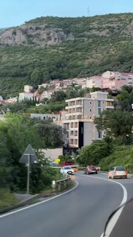 The video shows a winding road with several cars driving. On the left side of the road, there is a hotel with a sign that says "HOTEL". The hotel is a modern building with several floors and balconies. Behind the hotel, there are houses and buildings on a hillside covered with green trees. The sky is a pale blue, suggesting it is either early morning or late evening. The overall impression is of a scenic road leading to a hotel in a mountainous, coastal area.