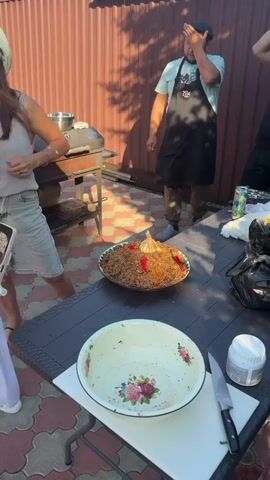 The video shows an outdoor scene with several people and food. A large platter of pilaf, garnished with garlic and chili peppers, is prominently displayed on a table. A man in an apron stands near a grill, appearing to be cooking or overseeing the food preparation. Another person is partially visible on the right side of the frame. The environment is a backyard with a wooden fence and paving stones. The lighting suggests it is daytime.
