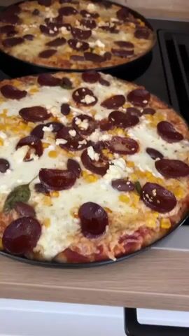 The video shows two pizzas on a kitchen counter. The pizza in the foreground is closer and in focus, while the pizza in the background is slightly out of focus. Both pizzas appear to be round and topped with cheese, sliced pepperoni or sausage, corn, and possibly some white cheese like feta. The background shows a wooden surface and a black stovetop. The lighting is natural and bright, suggesting it's daytime. The camera is held at an angle, focusing on the pizzas. There are no people visible in the video.