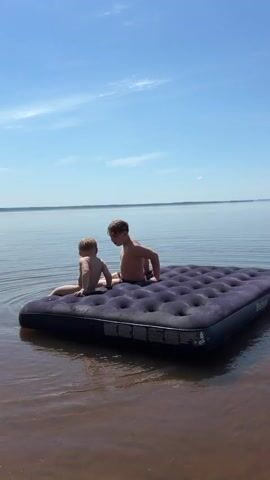 Two young boys are sitting on a dark blue inflatable mattress in a body of water. The sky is blue with a few scattered clouds. The water is calm, with a reddish-brown tint near the shore and appearing blue further out. The boys are both shirtless. The younger boy is in the front, facing forward and sitting up, while the older boy is behind him, sitting cross-legged. They appear to be interacting with the water.
