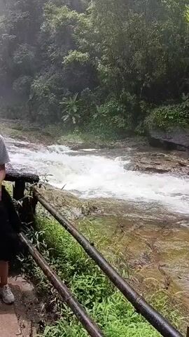 A woman and a young girl stand together on a path next to a railing. Behind them, a waterfall cascades down a rocky incline surrounded by lush green vegetation and trees. The woman, wearing a gray t-shirt and a light-colored crossbody bag, has her arm around the girl. The girl, dressed in a black hooded jacket and pink socks, leans her head on the woman's shoulder and smiles at the camera. The scene appears to be a natural outdoor setting with mist rising from the waterfall.