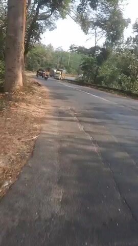 The video shows a road with a few vehicles. On the left side of the road, there are trees and a dirt embankment. The road itself is asphalt and appears to be in fair condition, with some cracks visible. In the distance, there are more trees and some structures. The sky is visible at the top of the frame and appears to be clear. There are at least two motorcycles and one truck visible on the road. One motorcycle is in the foreground, and another is further down the road. The truck is also further down the road. The video is shaky, suggesting it was filmed from a moving vehicle or by hand.