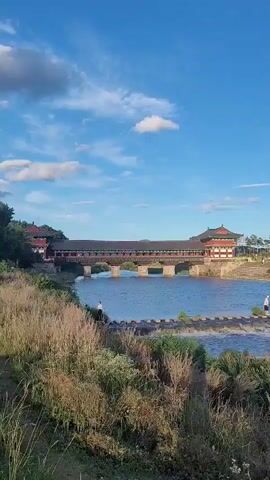 The video opens with a scenic view of a river with a traditional wooden bridge spanning across it. The bridge has red accents and a pagoda-style roof. In the foreground, tall grass grows along the riverbank. Several people are visible, including two individuals standing on stepping stones in the river and a person walking on the stones further to the left. On the far bank, behind the bridge, there's a grassy area with trees and a hillside. The sky is blue with a few scattered white clouds. The lighting suggests it's daytime, likely late afternoon or early evening due to the warm tones in the grass.