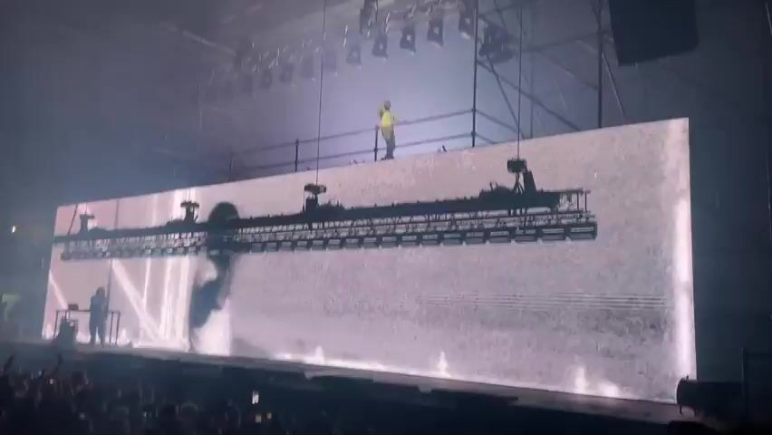 A concert is taking place inside of a large venue filled with people. A man in a yellow shirt stands on a raised platform atop of large screen facing the crowd. The screen displays a video of the same man, but wearing a white shirt and holding a microphone. Bright white lights shine down on the stage, and scaffolding surrounds the set. A person can be seen standing behind the screen to the left, near the floor.