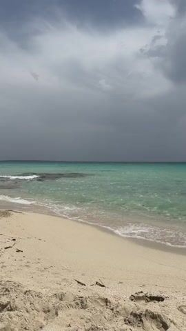 A serene beach scene with gentle waves lapping at the shore under a dramatic, cloudy sky. The sand is light beige and shows subtle textures. The water is a clear turquoise, shimmering with sunlight filtering through the clouds. In the distance, the horizon is marked by a darker band of water or land, with the ominous, dark gray clouds filling the upper portion of the frame. There are no people or man-made objects visible in the frame, creating a sense of peaceful solitude.