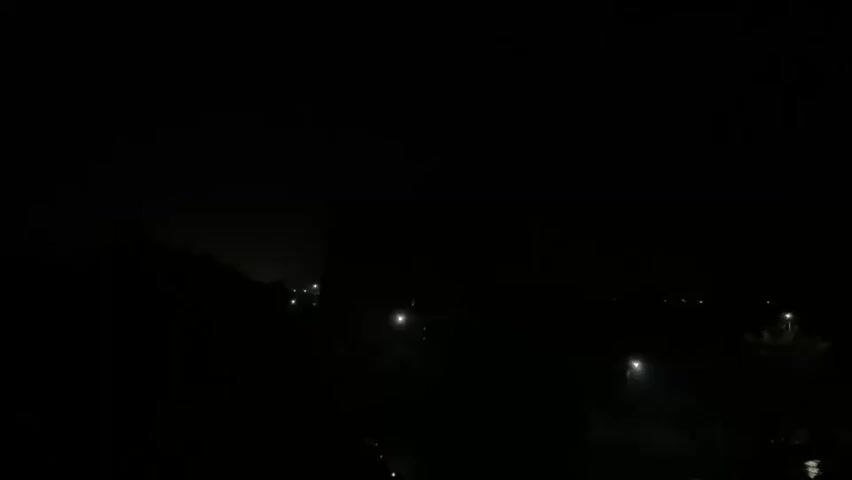 The video shows a dark, nighttime scene with a few streetlights visible in the distance. The sky is dark and appears to be cloudy or foggy. There is a faint glow in the sky, suggesting distant city lights or possibly lightning. The overall impression is one of a quiet, somewhat eerie, nocturnal environment.