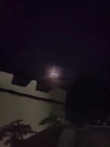 The video shows a night sky with a bright light in the distance. The light appears to be moving. The sky is dark and there are no stars visible. There is some blurry foliage in the foreground.