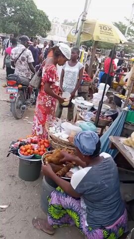A short video captures a bustling scene at what appears to be a marketplace. Several people are visible, some interacting with vendors. One woman, wearing a colorful red floral dress, is seen purchasing items from a stall. Another woman, seated on a barrel, is handling and sorting what looks like brown root vegetables. To her left, there is a bucket filled with vibrant red tomatoes. A motorcycle is parked nearby. Overhead, an umbrella provides shade to one of the stalls. The area is unpaved and appears to be somewhat sandy or dusty. The scene is filled with activity and various goods for sale, suggesting a lively commercial setting.