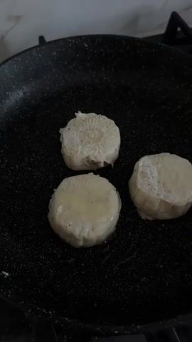 Three round, flat food items are being fried in a black nonstick skillet on a stovetop. The items appear to be small cakes or pastries, with decorative patterns pressed into their tops. One item on the right is being touched with a fork, which is lifting its edge slightly. The food is pale in color, with some parts beginning to brown from frying. The skillet has a speckled surface and is on a dark stovetop. The background is out of focus and appears to be a kitchen environment.