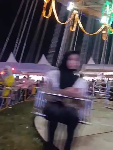 A boy in a blue shirt and jeans rides a swing carousel at night. The carousel is decorated with colorful lights, including strings of orange and green lights above and a large, illuminated circular design in the background. Other people are visible on the ride and in the background, near tents and other fair attractions. The carousel is in motion, causing some of the figures to appear blurred.