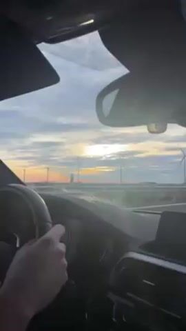The video, taken from inside a moving car, shows a field of wind turbines at sunrise. The sky is cloudy, with the sun partially visible behind the clouds, creating a warm orange glow on the horizon. The driver’s hand is visible on the steering wheel. The car is on a highway, separated from the wind turbines by a guardrail. Another car passes in the opposite direction. The camera angle is slightly obscured by the car's interior.