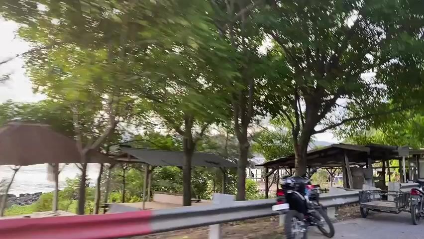 The video captures a roadside scene with several motorcycles and a black SUV parked. There are small open-air shelters with corrugated roofs, possibly for vendors or rest stops, located near a body of water. Lush green trees with dense foliage line the road, and the sky appears overcast. In the distance, islands or hills can be seen across the water. The camera appears to be moving, suggesting it is filmed from a vehicle.