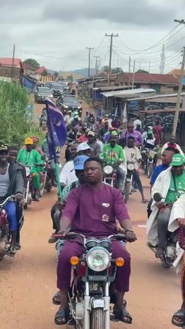 The video shows a procession of motorcycles and a few cars traveling down a dirt road lined with buildings and vegetation. The lead motorcycle is ridden by a man in a purple tunic and pants, with another person behind him. He is looking straight ahead. Several other motorcycles follow, some carrying two people. Many individuals in the procession are wearing green shirts and hats, and some are holding purple flags with white lettering. In the distance, a line of cars, including a police car with flashing lights, is visible. The sky is overcast. The overall atmosphere appears to be a political rally or a motorcade.