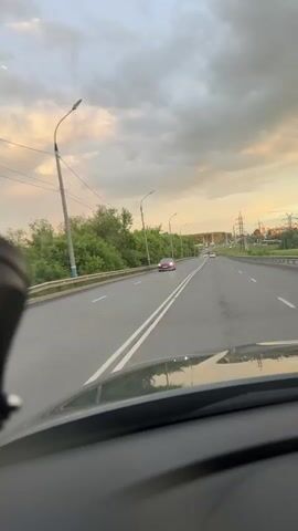 This video captures a driving POV shot on a highway in Arzamas, Russia. The perspective is from inside a vehicle, looking out through the windshield at the road ahead and the sky.

The primary subject is the highway itself, which appears to be a multi-lane road with clear lane markings. Several cars are visible on the road, traveling in the same direction as the viewer. One car, a reddish-pink sedan, is in the left lane ahead. Further down the road, other vehicles are spaced out. To the left of the highway, there is a lush green tree line bordering the road, with a metal guardrail separating the road from the trees. Streetlights are positioned along the side of the highway.

The setting is an outdoor, daytime scene. The time of day appears to be late afternoon or early evening, indicated by the warm, soft lighting and the colors in the sky. The sky is partially cloudy, with dramatic, textured clouds that are illuminated by the setting sun, casting a golden and pinkish hue. The overall mood is peaceful and tranquil, with the gentle movement of the car contributing to a sense of journey.

There are no visible people in the immediate foreground or as distinct subjects. The focus is on the road, the traffic, and the natural environment under a dynamic sky. Power lines and utility poles are visible in the distance on the right side of the highway, suggesting an urban or suburban fringe area. The asphalt of the road appears wet, possibly from recent rain, which adds a reflective quality to the surface. No text is visible in the video.