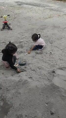 Two young girls are playing in a sandy area. One girl is wearing a pink shirt and white shorts, and the other is wearing a black shirt and a pink skirt. They both have shovels and are digging in the sand. There is a small tricycle in the background. The environment is outdoors, with dirt and vegetation visible. The sky is overcast.