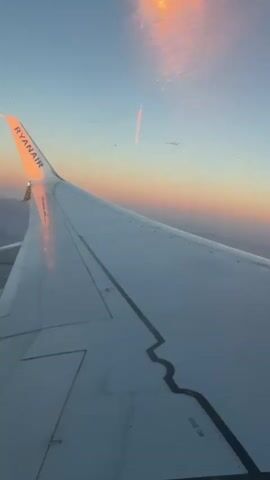 The video shows a view from an airplane window. The right wing of the plane, with the Ryanair logo visible on the wingtip, fills the foreground. The wing is white with black markings. Outside the window, the sky is a gradient of colors from light blue at the top to pink and orange near the horizon, indicating either sunrise or sunset. Clouds are visible in the distance, catching the warm light. The camera is stationary, capturing a static view from the aircraft.