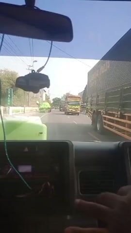 The video captures a point-of-view shot from inside a vehicle, looking forward through the windshield. The scene is a two-lane road with moderate traffic. On the right side of the road, buildings and some construction materials, possibly concrete pipes, are visible. Several trucks are on the road, some carrying large loads covered with green tarps. A motorcycle is also seen. The time of day appears to be daytime, with bright sunlight. The interior of the vehicle shows a dashboard and a hand, presumably the driver's, on the steering wheel. The overall impression is that of a typical road scene in an urban or semi-urban environment.