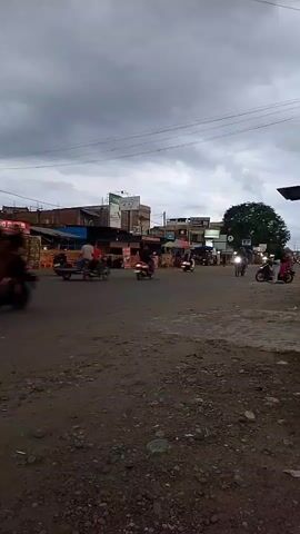 The video shows a street scene with several motorcycles and a three-wheeled vehicle. The road appears to be unpaved or partially paved. There are buildings on the left side of the road, some of which appear to be shops or market stalls. The sky is overcast, suggesting cloudy or stormy weather. The overall atmosphere is that of a busy street in a town or city, likely in a developing country, given the types of vehicles and the informal nature of some of the businesses.