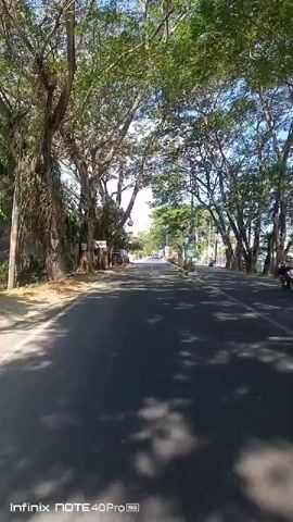 A point-of-view video shot from a moving vehicle shows a road lined with trees on both sides. Sunlight filters through the leaves, casting shadows on the asphalt. On the left side of the road, there are some buildings and parked vehicles. On the right, there is a sidewalk with a fence and some smaller trees. The road leads into the distance, where more trees and buildings are visible. The sky is blue with some scattered clouds. The video is watermarked with "Infinix NOTE 40 Pro 5G".