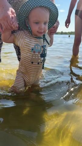 A baby wearing a striped onesie and a green polka-dot hat stands in shallow water, held by an adult's hands. The baby's feet are submerged in the water, which is clear with sandy bottom visible. Other people are in the background, some swimming and others relaxing on an inflatable raft. The sun is shining, casting a glare on the water's surface.