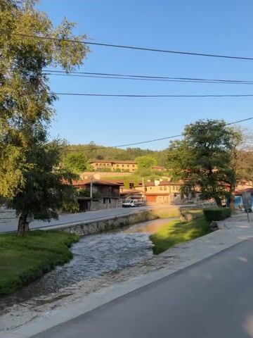 This video showcases the picturesque town of Koprivshtitsa, Bulgaria, during what appears to be late afternoon or early evening. The setting is a charming, historic town with traditional Bulgarian architecture, characterized by colorful houses with wooden accents and tiled roofs. A clear blue sky suggests good weather.

The video begins with a view of a small river or stream flowing through the town, with green grassy banks and stone-lined sides. Traditional houses and trees line the banks. As the camera pans, it reveals more of the town's unique architectural style, including a yellow house with a stone foundation and a sign in front of it. A prominent utility pole with numerous wires is visible in this shot.

Further on, the scene shifts to a park-like area with cobblestone pathways and lush green grass. Several tall evergreen trees provide shade, and the sunlight filters through their branches, creating a warm glow. A stone monument, possibly a memorial, stands in the center of the grassy area, flanked by smaller stone sculptures. The overall atmosphere is peaceful and serene, evoking a sense of historical charm and natural beauty.

No people are visible in the video. The activity is a slow, panning tour of the town's streets, river, and park. The visible text on the sign in the second clip is partially obscured but appears to be a restaurant or guesthouse name, with a phone number below it. The time of day is suggested by the long shadows and the warm, golden light illuminating the trees and buildings. The location cues, such as the architectural style and the monument, are consistent with Koprivshtitsa.