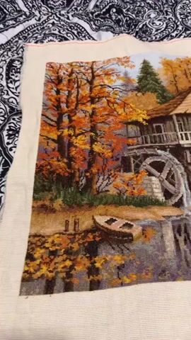 The video displays a close-up of an unfinished cross-stitch tapestry depicting an autumnal scene. The tapestry features a water mill with a large wheel, a stone and wooden building, trees with vibrant orange and yellow foliage, a small boat, and a body of water reflecting the surrounding colors. The tapestry is laid out flat, with the edges showing the unstitched canvas. The camera slowly pans across the tapestry, highlighting the detailed stitching and the rich colors of the autumn landscape.