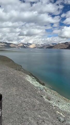A vertical video pans across a wide, serene, turquoise lake. The lake is surrounded by barren, mountainous terrain. The sky is partly cloudy with dramatic white clouds against a bright blue backdrop. The foreground consists of a rocky, gravelly slope leading down to the water's edge. There is a solitary utility pole visible on the lakeshore in the mid-ground of the video. The overall scene evokes a sense of vastness and natural beauty.